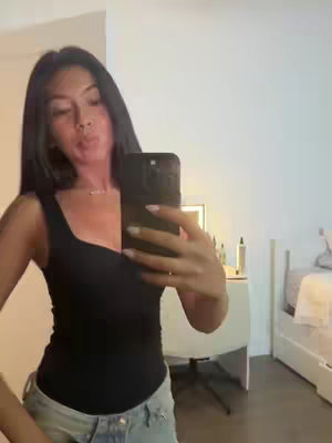 A selfie-style shot of a young woman in her room. She is holding a phone up, appearing to take the photo, with a slight pout. She is wearing a black tank top and light-colored jeans. The background includes a white desk and part of a bed.