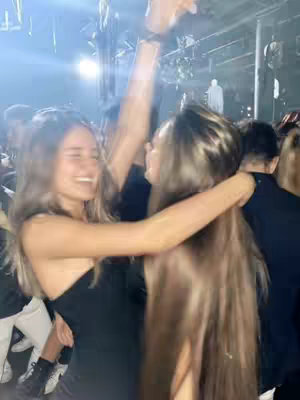 Two young women are embracing and laughing, likely at a party or event, with their arms wrapped around each other. The background is blurred, suggesting motion, and filled with lights and other people. The woman in the foreground is wearing a black dress and smiling, while the other is partially obscured by her long hair.
