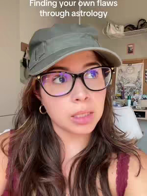 A woman wearing glasses and a cap is looking at the camera and talking, with the text overlayed at the top. The background is a room with a white wall. The overall composition is a selfie-style shot.