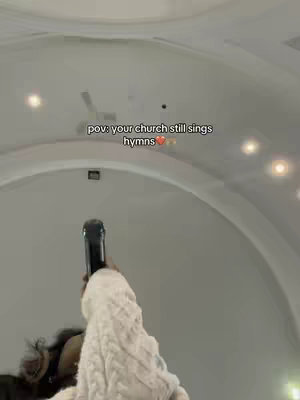 The cover image shows a person holding a microphone, singing in what seems to be a church setting. The person is wearing a white knitted sweater, and their face is partially visible, looking up towards the ceiling. The ceiling is high and vaulted, with a few lights visible. Text overlay at the top reads, 'pov: your church still sings hymns'. The overall composition suggests a live performance or worship scene.