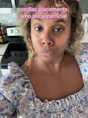 A woman with curly blonde hair, wearing a floral top, is centered in the frame. The background kitchen setting is slightly visible. Text overlayed in a bubble font states, 'zodiac placements who are superficial'. The woman has a serious expression.