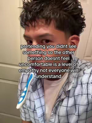 A selfie-style shot featuring a young man in a plaid shirt. The man is casually styled with short, dark hair. He's positioning himself in front of what appears to be a bathroom mirror, with a toothbrush casually placed in his mouth. The overall composition is intimate, given that the camera is close to the man.