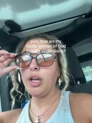 A selfie-style video showing a young woman inside what appears to be a car. She's wearing eyeglasses and a light-colored tank top. The background is somewhat blurred but hints at an outdoor setting. The overall lighting is natural.