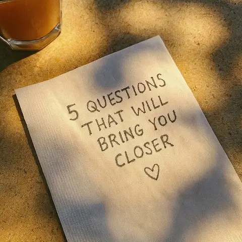 A close-up shot of a white napkin with handwritten text and a coffee cup in the upper left corner. The napkin has the text, '5 QUESTIONS THAT WILL BRING YOU CLOSER' with a heart drawn after the word closer. The background is out of focus, suggesting a table surface. The lighting creates shadows making the text more readable.