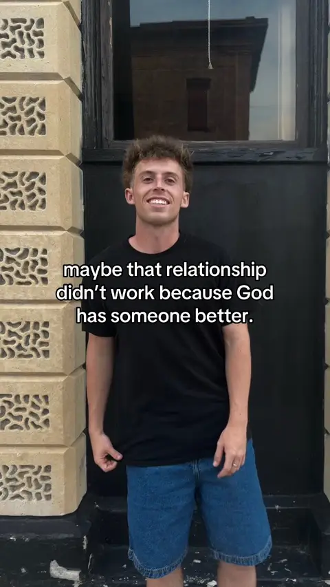 The image shows a young man standing outdoors, leaning near a building facade with a window. He's smiling and dressed in a black t-shirt and denim shorts. Text is overlaid, suggesting a relationship-related message.
