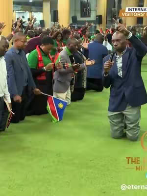 The cover image shows a large group of people, predominantly men, kneeling in prayer on a green floor. A figure in a blazer stands at the front, holding a microphone and pointing upwards. Behind him, people have their hands raised and flags, including a Namibia flag. The background is an interior setting.