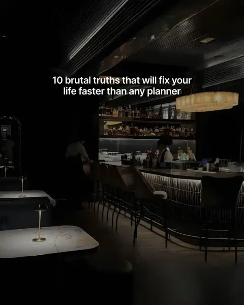 A dimly lit interior shot of a bar or restaurant, with black walls and dark wooden accents. The scene is dominated by the bar counter, backlit with a line of bottles and glassware, and several bar stools.
