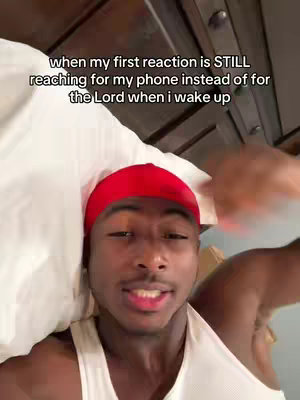 A selfie-style shot of a young man lying in bed. He is wearing a white tank top and a red head covering and is looking at the camera with what appears to be a serious or introspective expression of the face.