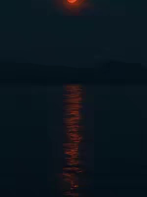 A dark, moody image of a body of water with a reflection of the sunset. The sky is a dark blue, with a large orange sun near the horizon line, suggesting the sky is still dark. The water reflects the orange light from the sun in a long, vertical streak, creating the illusion of fire. The scene is dim with the dark night.