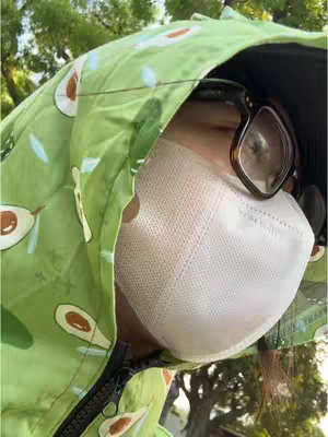 The cover image is a selfie of a person from the chest up, shot outdoors on a sunny day. The person is wearing sunglasses and a light green, hooded jacket with an avocado pattern. A white face mask covers their mouth and nose. The background suggests a park setting with trees.