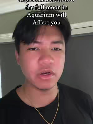 A vertical video cover features a close-up of a young man's face, framed from the chest up. He has dark hair, fair skin, and is wearing a black shirt with a gold chain necklace. The background is blurred, suggesting an interior setting with neutral-colored walls. Text overlay in the upper half of the frame reads, 'Capricorn here's how the full moon in Aquarius will Affect you'. The man's expression is neutral, looking directly at the camera with his mouth slightly open.