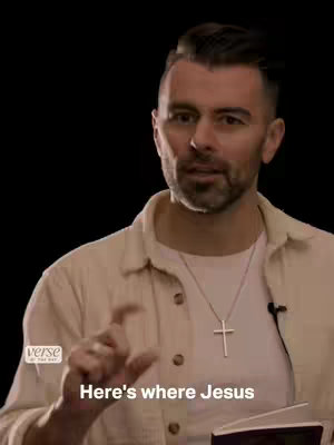 A man with short dark hair and a beard is centered in front of a black background, giving a speech. He wears a cream-colored shirt and a cross necklace, and gestures with his right hand.