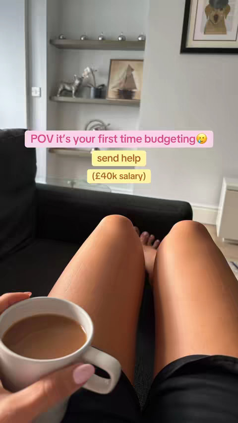 The image shows a person's legs, holding a white mug with a brown beverage, likely coffee. The person is seated on a black couch, with a shelf in the background. Text overlays on the photo read "POV it's your first time budgeting 🥺 send help (£40k salary)".