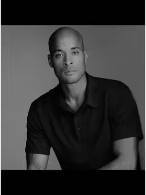 A black and white portrait featuring a bald man in a black shirt looking towards the camera. The background is plain and light-toned. The man's features are well-defined, and the lighting creates contrast.