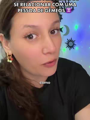 A woman with brown hair and fair skin looks directly at the camera. She wears gold hoop earrings with colorful charms. The background is out of focus, with some decorations and text that are part of the video's overlay.
