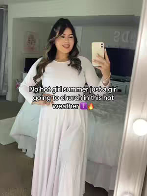 A woman wearing a long white dress, holding a phone up to take a mirror selfie. The background shows elements of a bedroom, with items such as a bed and a framed picture on the wall. Text overlayed on the image reads, "No hot girl summer just a girl going to church in this hot weather".
