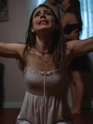A woman in a white dress is kneeling with her arms outstretched, eyes looking upward with an expression of distress. Additional figures appear blurred in the background, suggesting a scene with multiple figures or a dynamic event. The overall composition is softly lit.