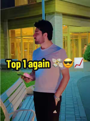 A man in a white t-shirt and black pants is standing outside at night, holding a phone and looking to his left. The cover image is overlaid with text and graphics. The words "Top 1 again" are placed in a yellow box in the bottom left corner, and to the right of the man's head are several emojis: money with wings, sunglasses, and an upward-pointing red graph. The background includes greenery, a building with a glass entrance, and other architectural details.