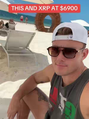 A man in sunglasses and a white cap is sitting on a beach with a blurred beach scene in the background. Text overlayed in a red bar at the top states, "THIS AND XRP AT $6900"