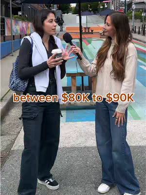 Two women stand on a city street, one holding a microphone and interviewing the other. The interviewee gestures with one hand while holding a coffee cup. Text on the image indicates a salary range. The background includes buildings, an umbrella, and partially visible city artwork.