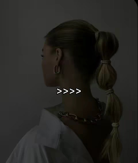 The image shows a woman from the back, with her hair styled into a high ponytail with bubble sections. She's wearing a white shirt and a gold chain necklace, and her profile is visible against a neutral background.
