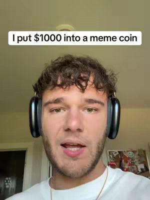 A young man with short, curly brown hair is facing the camera, wearing large black headphones. He has a light complexion and a slight beard, with a white t-shirt. Above him, in a white rectangular box, the text overlay reads, 'I put $1000 into a meme coin.' The background appears to be a room with a pale wall.