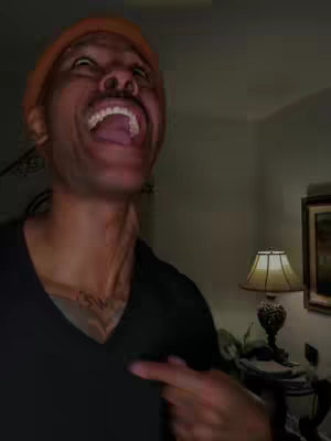 A man with a wide open mouth, eyes looking upwards, pointing at his neck. He is wearing an orange knit cap and a black shirt. The background shows a room with a lamp and a picture.