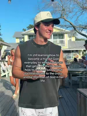 A young man wearing a casual outfit stands on a wooden deck with several buildings in the background. He is smiling with some text overlaid on the image.