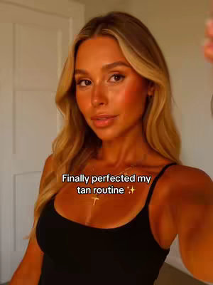 A woman with long blonde hair and a bronzed complexion poses for a selfie. She is wearing a black tank top and a delicate gold necklace. The background is minimalistic, with a neutral-colored door or wall that complements her warm skin tone. The lighting is soft and flattering, highlighting her features.