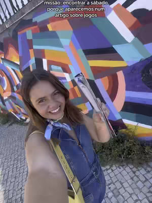 A selfie-style shot with a woman holding a magazine, smiling at the camera, with a colorful, abstract mural in the background.