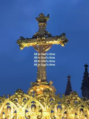 A vertical shot of a golden cross illuminated against a deep blue sky. The cross is ornate, with intricate detailing, and stands atop a structure with similar golden filigree. Below the cross, a repeating text overlay declares 'All in God's time,' adding a message of faith and patience.