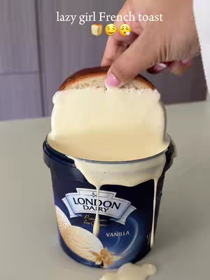 A close-up shot showing a slice of bread being dipped into a container of vanilla ice cream, with the ice cream overflowing. The background is blurred, keeping the focus on the food and hand. The branding of the ice cream is clearly visible.