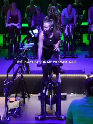 The image captures a woman leading an indoor cycling class. She is centered on an exercise bike, facing forward with an energetic expression. Other class participants are visible in the background, all riding on stationary bikes, in a studio lit with purple and green lights. White text overlays at the bottom of the image reads "THE PLAYLIST FOR MY WORSHIP RIDE".