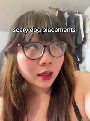 A close-up shot of a woman with long brown and blonde hair, wearing black-rimmed glasses and red lipstick. She is looking up and to the right with a slight expression of surprise or thought. The background is blurred, suggesting a casual indoor setting with clothing hanging on a rack.