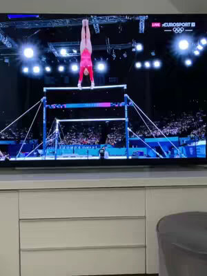 A large flatscreen TV displays a live sports broadcast of a gymnastics competition. The gymnast is performing a handstand on uneven bars. The bottom part of the image shows a light-colored cabinet with drawers, positioned beneath the TV. A blurred gray object, possibly a chair or the side of a sofa, is visible below.