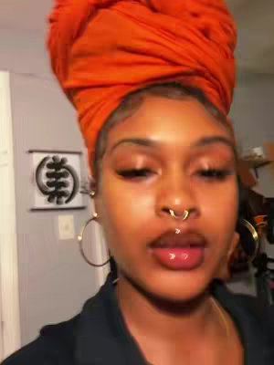 A close-up shot features a woman of color wearing an orange head wrap. She has full lips, a nose ring, and large hoop earrings. The background is a room with some decorative elements, including a framed symbol on the wall.