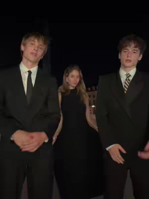 Three people are featured in the image against a dark background. Two individuals, assumed to be men, are standing on either side of a woman, all dressed in formal black suits and dresses suggesting a gala or formal event. The lighting is dim, focusing attention on the individuals.
