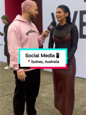 A man wearing a pink hoodie interviews a woman holding a microphone. A graphic with text and location tag on the bottom.