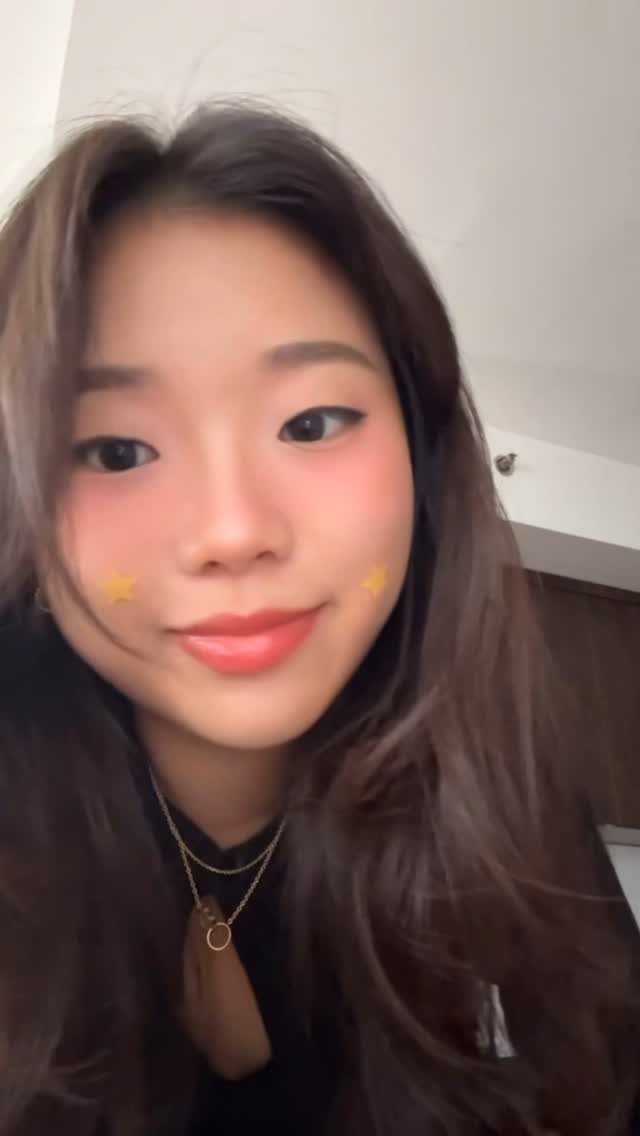 A close-up selfie of a person with fair skin, long brown hair, and blush applied to their cheeks. They are wearing a black top with several gold necklaces and a gold ring. There are two small yellow star stickers on their cheeks. The background is a plain wall.