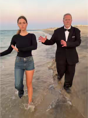 A woman in casual clothes and a man in a tuxedo stroll together through shallow ocean water, with a beach and sunset sky in the background. They are both extending their arms out to the sides.