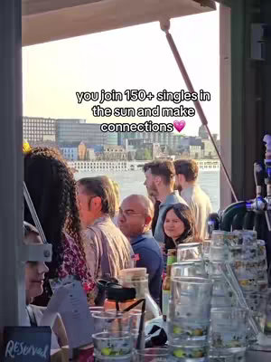 An outdoor scene on a sunny day, likely a gathering or event. People are standing around a bar or counter visible on the right side and there is a body of water and buildings in the background. A text overlay says 'you join 150+ singles in the sun and make connections'
