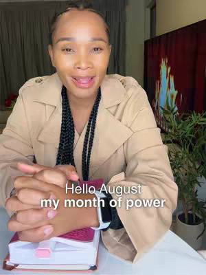A woman with dark braided hair is speaking, wearing a beige trench coat, and resting her hands on a pink book. Text overlaid reads 'Hello August my month of power'. The background includes a green plant and a blurry TV.