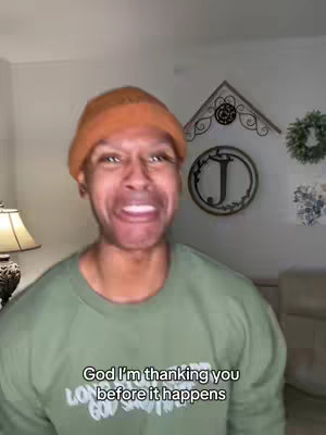 A man with an orange beanie and green shirt makes a comical, expressive face, appearing to be ecstatic. Text at the bottom reads, 'God I'm thanking you before it happens.' The background consists of a plain wall with decorative items.