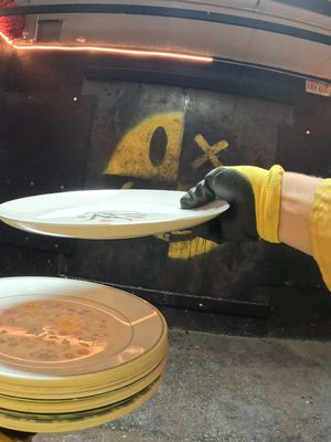 A person wearing a yellow-sleeved work glove holds a white plate in front of a dark background. Other plates are stacked below. The background shows a painted yellow design.