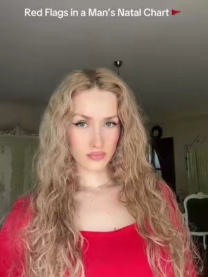 A woman appears in a selfie-style shot, gazing earnestly at the camera. She has long, wavy blonde hair framing her face, and her striking features include defined eyebrows, dramatic eyeliner, and a soft rosy lips. She is wearing a vibrant red top. The background is a neutral beige.