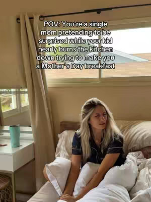 A woman with light hair and navy blue pajamas sits up in bed, looking to the side with a slightly concerned expression. She is propped against pillows with rumpled bedding. The background includes a window with blinds partially drawn, and a bedside table with a teal tumbler on top.