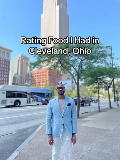 A man in a light blue blazer and white pants stands on a city street, with a blue bus and tall buildings in the background. Text overlayed on the image reads, 'Rating Food I Had in Cleveland, Ohio.'