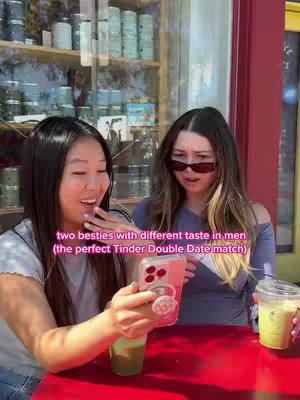 Two young women are seated at an outdoor table. The woman on the left, with long dark hair parted to the side, is smiling brightly at a phone she's holding. The phone has a pink case and is angled towards her and away from the camera. The other woman, with long brown hair and sunglasses, looks with a surprised or concerned expression. Both women are holding drinks. The background shows shelves and glass windows. The scene appears bright and shot in natural light.