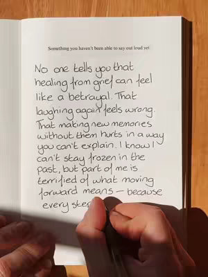 A hand holding a pen writes on an open notebook. The notebook is on a wooden surface. The text in the notebook is handwritten. The overall composition is a top-down view.