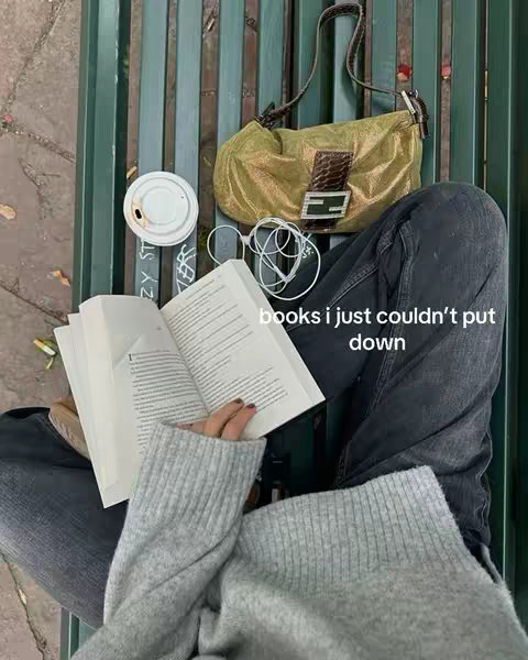 A person is sitting on a green bench with a book open in their lap. A grey sweater covers their upper torso and arms. In the background are a small gold shoulder bag, a cup of coffee with a white lid, and white earphones. The words “books i just couldn’t put down” are overlaid onto the image.